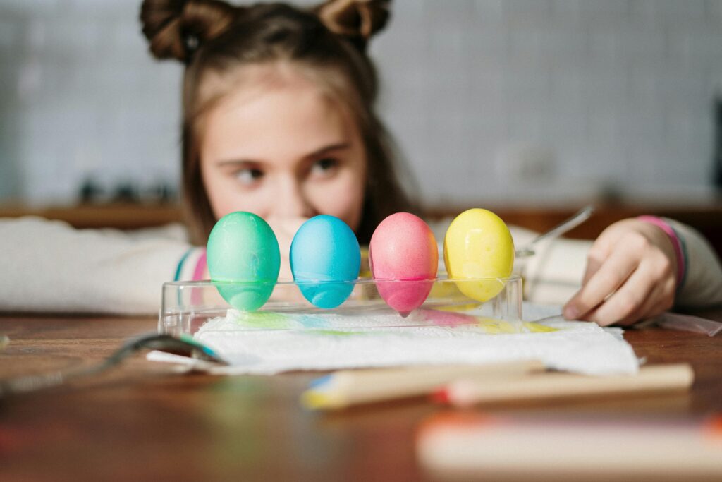 A child happily decorates colorful Easter eggs on a wooden table indoors.