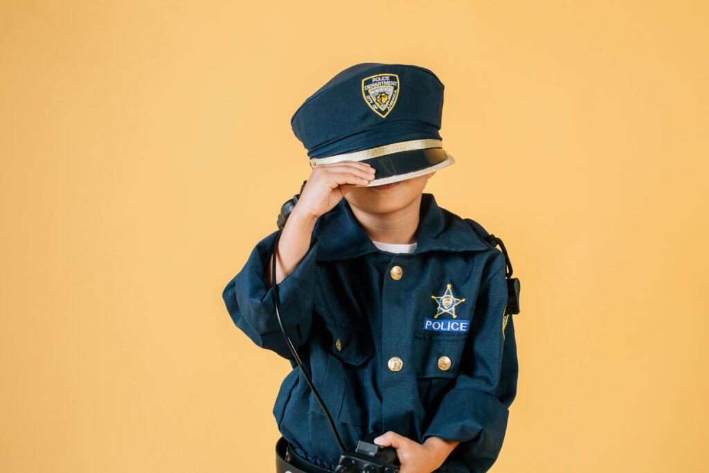 Unrecognizable child in police uniform standing in studio with transceiver in hand and pulling cap over face on yellow background